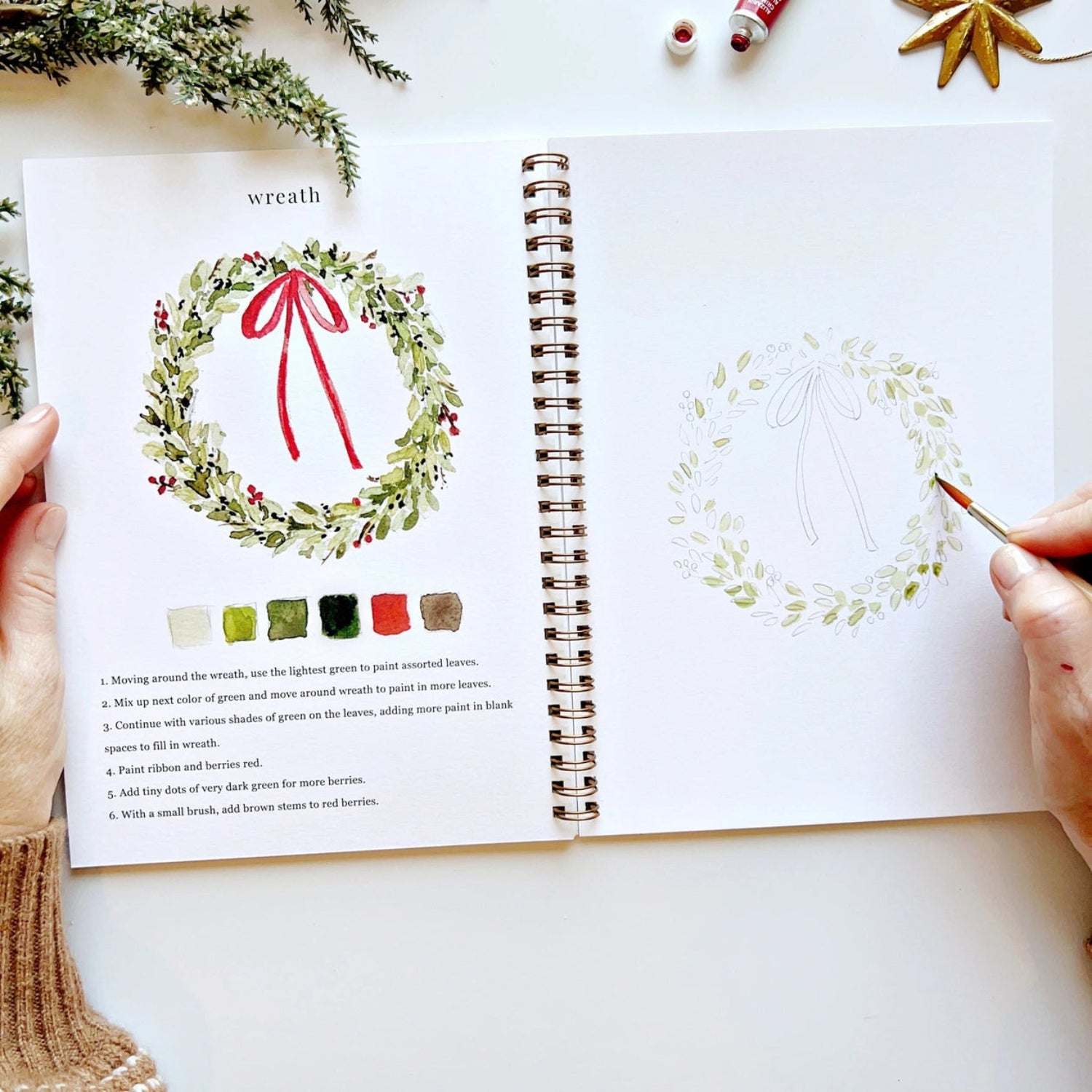 Open book with a Christmas wreath illustration and color guide on a white surface.