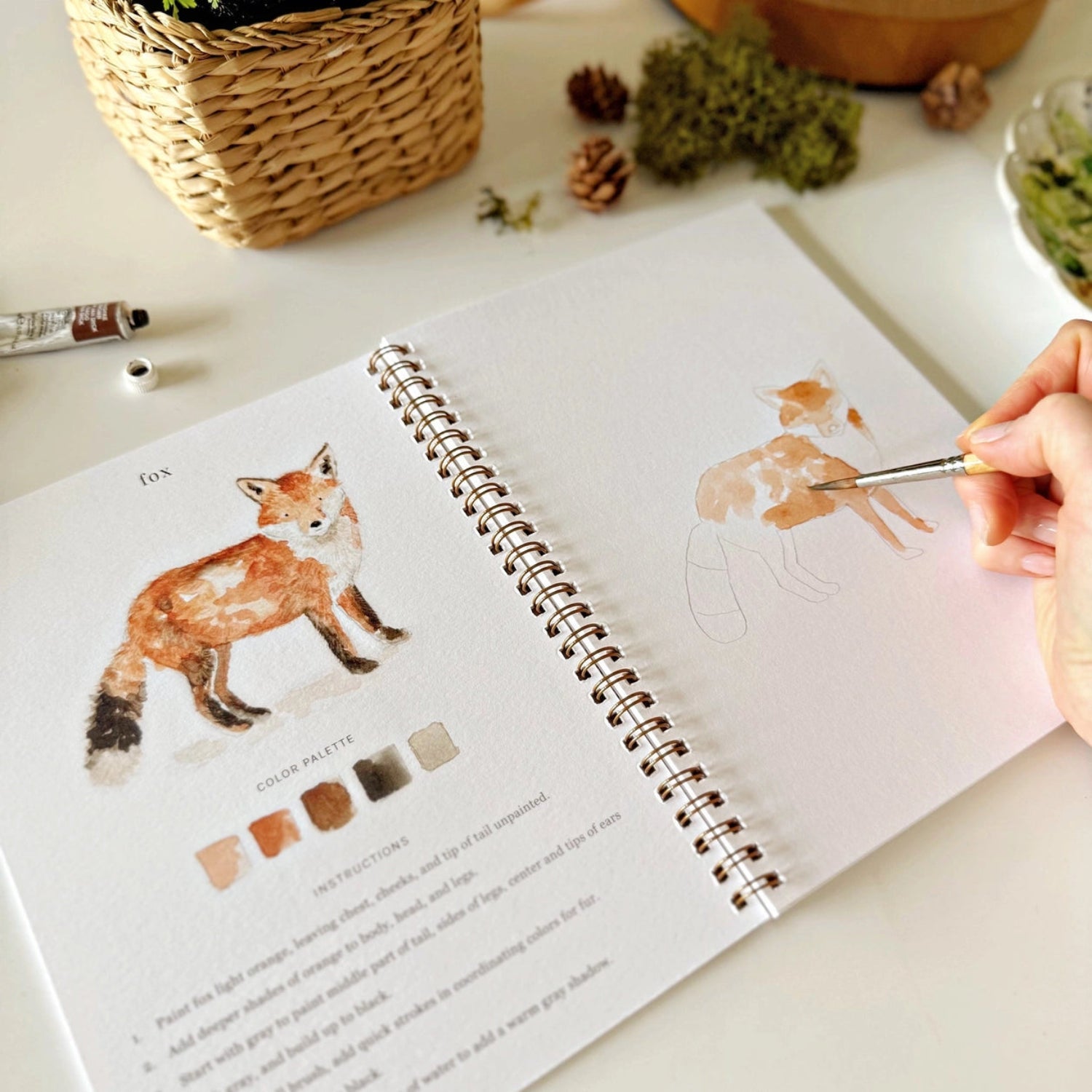 Person painting a fox illustration in an art book with a paintbrush.