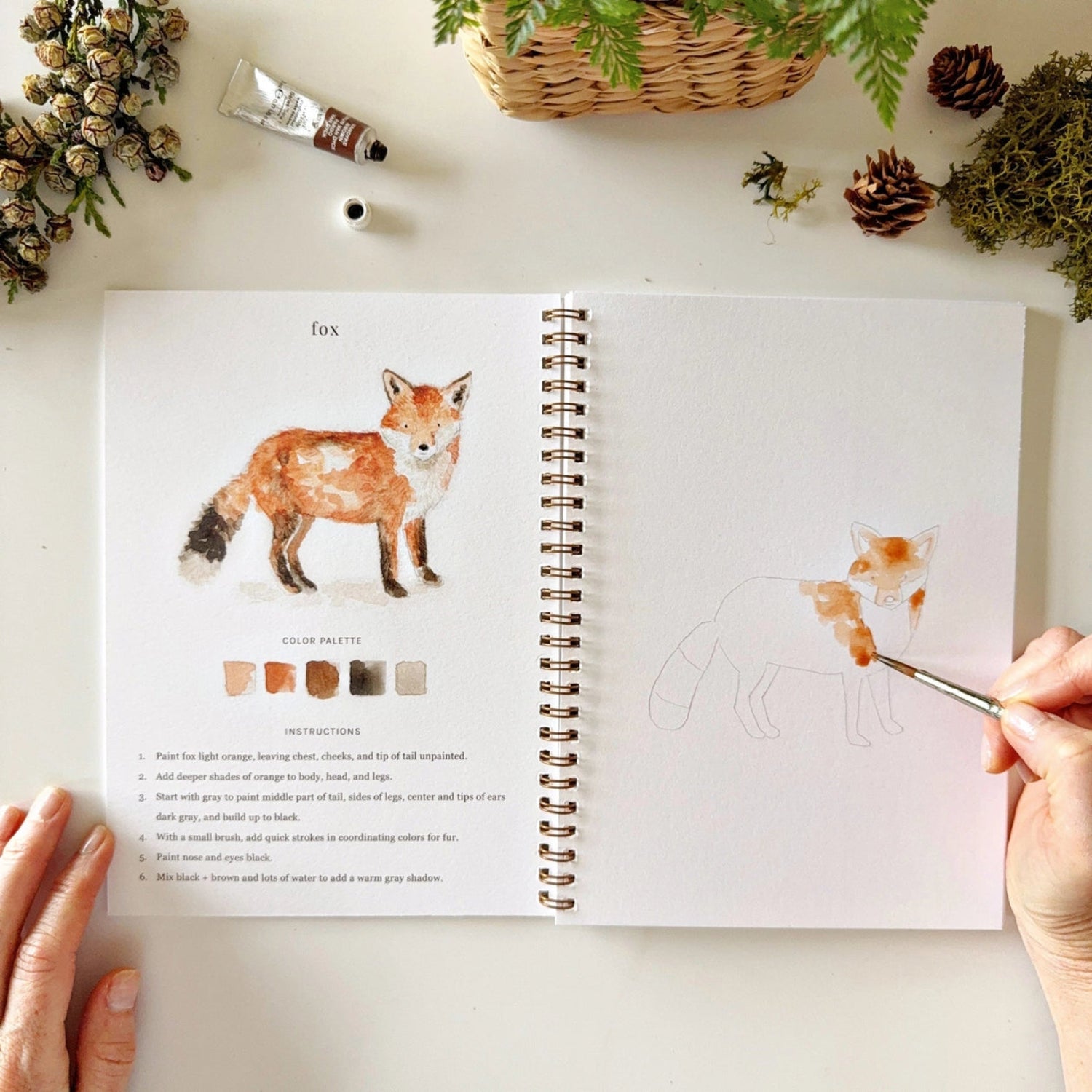Open book with a fox illustration and color chart, being painted by a hand with a brush.