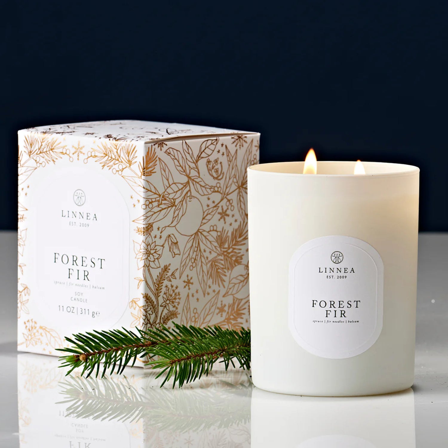 Linnea Forest Fir candle with packaging and a sprig of pine on a reflective surface.