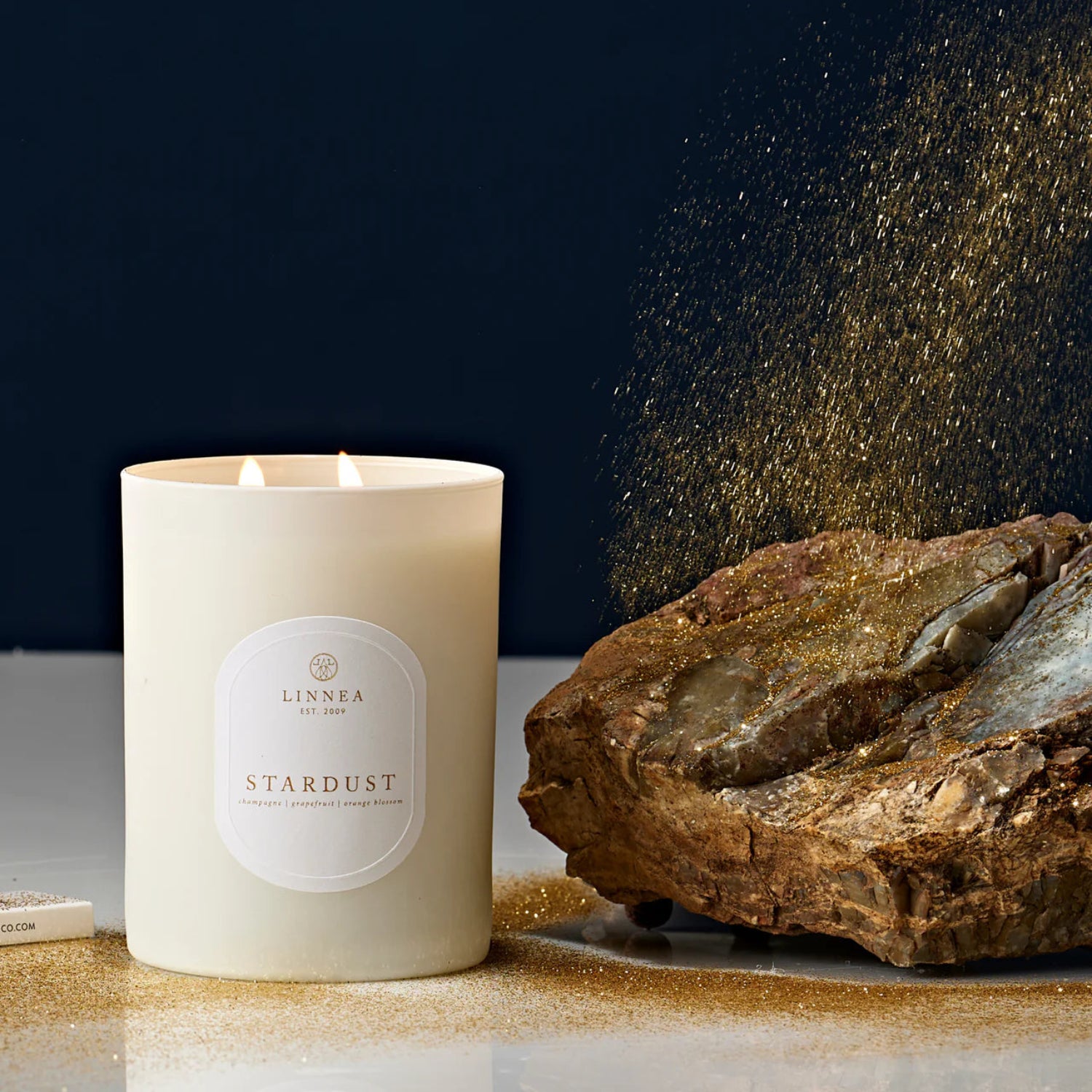 Candle labeled 'Stardust' by Linnea next to a rock with gold dust on a dark background
