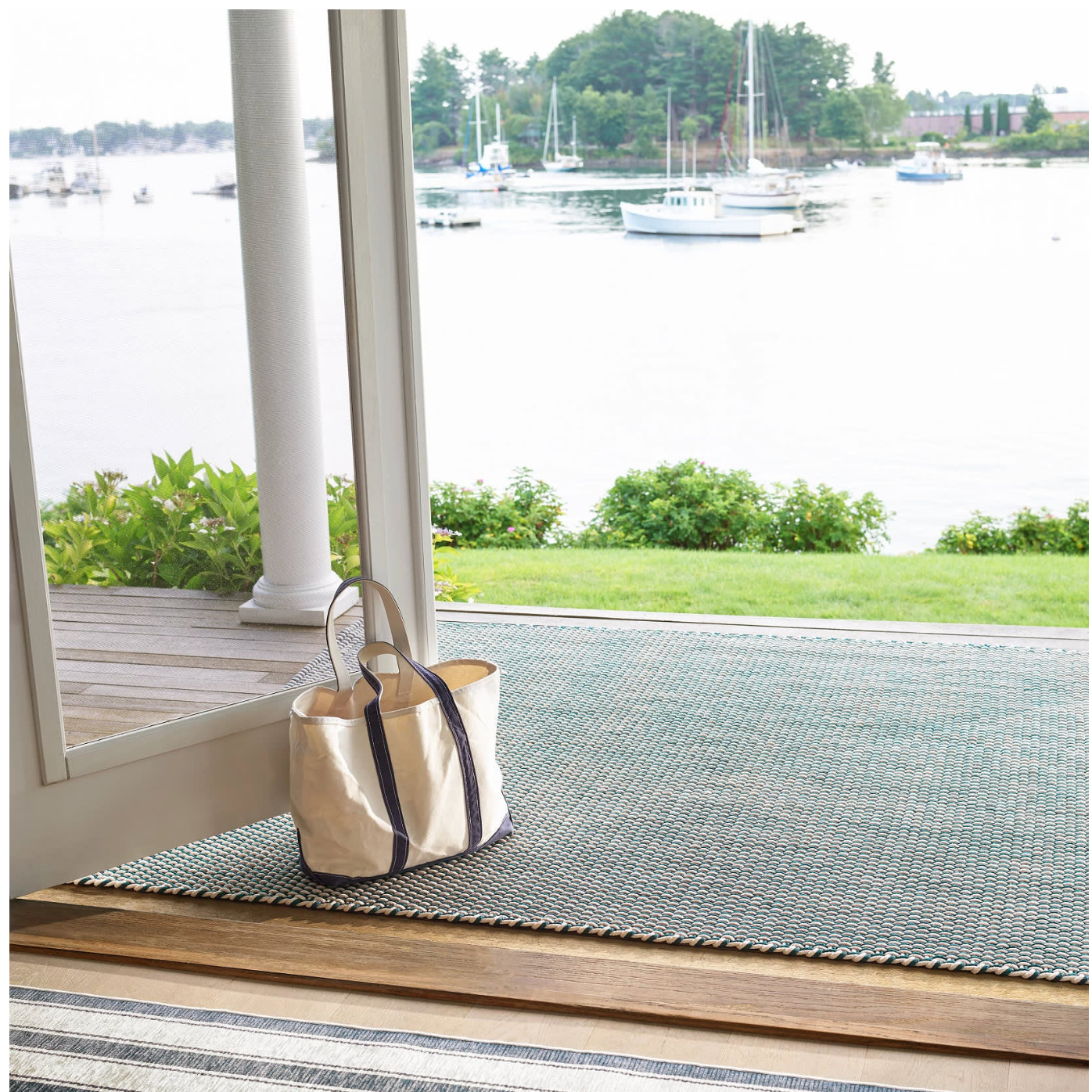 Tote bag on a striped rug with a view of a harbor through large windows.