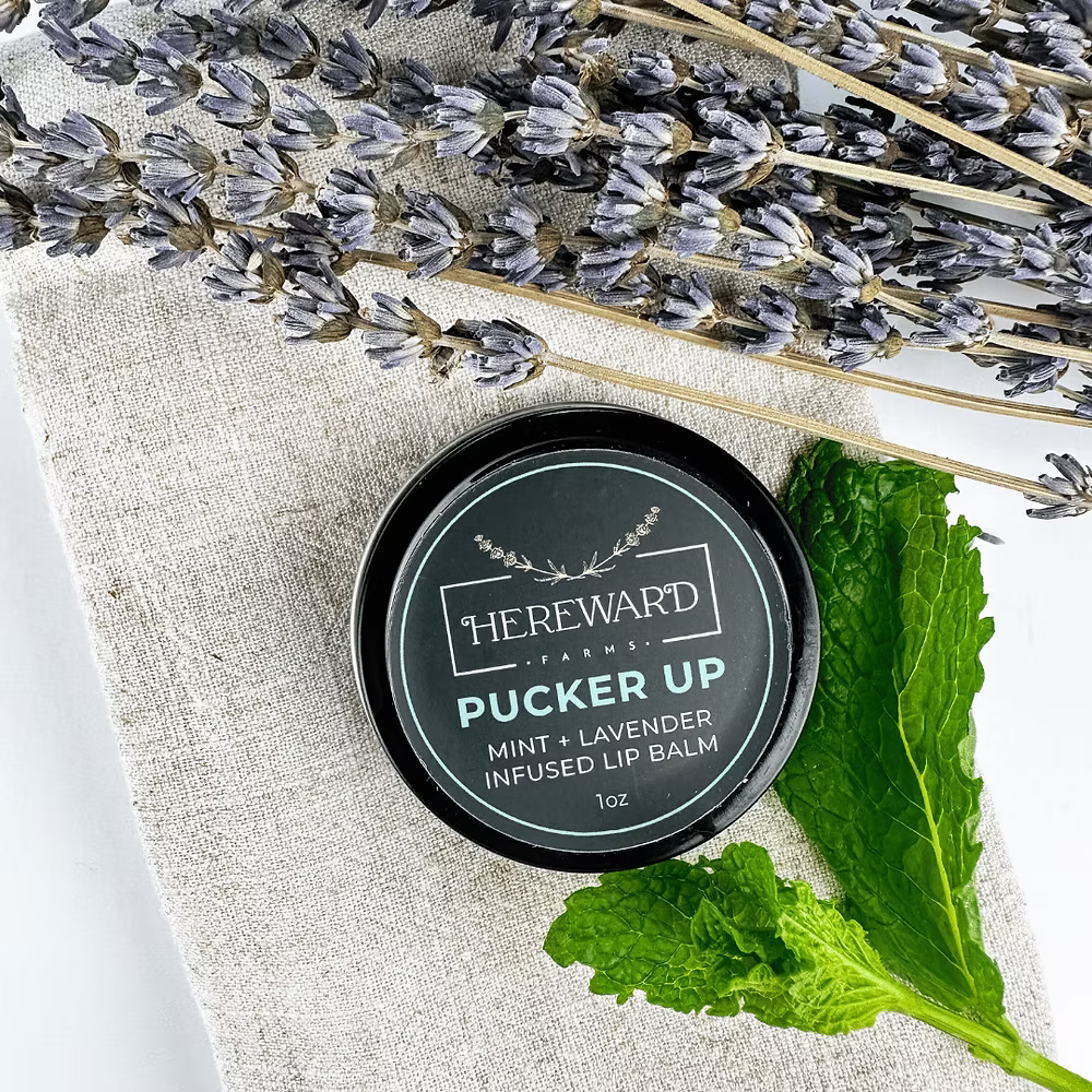 Lip balm container labeled 'Hereward Farms' with lavender and mint leaves on a textured fabric background