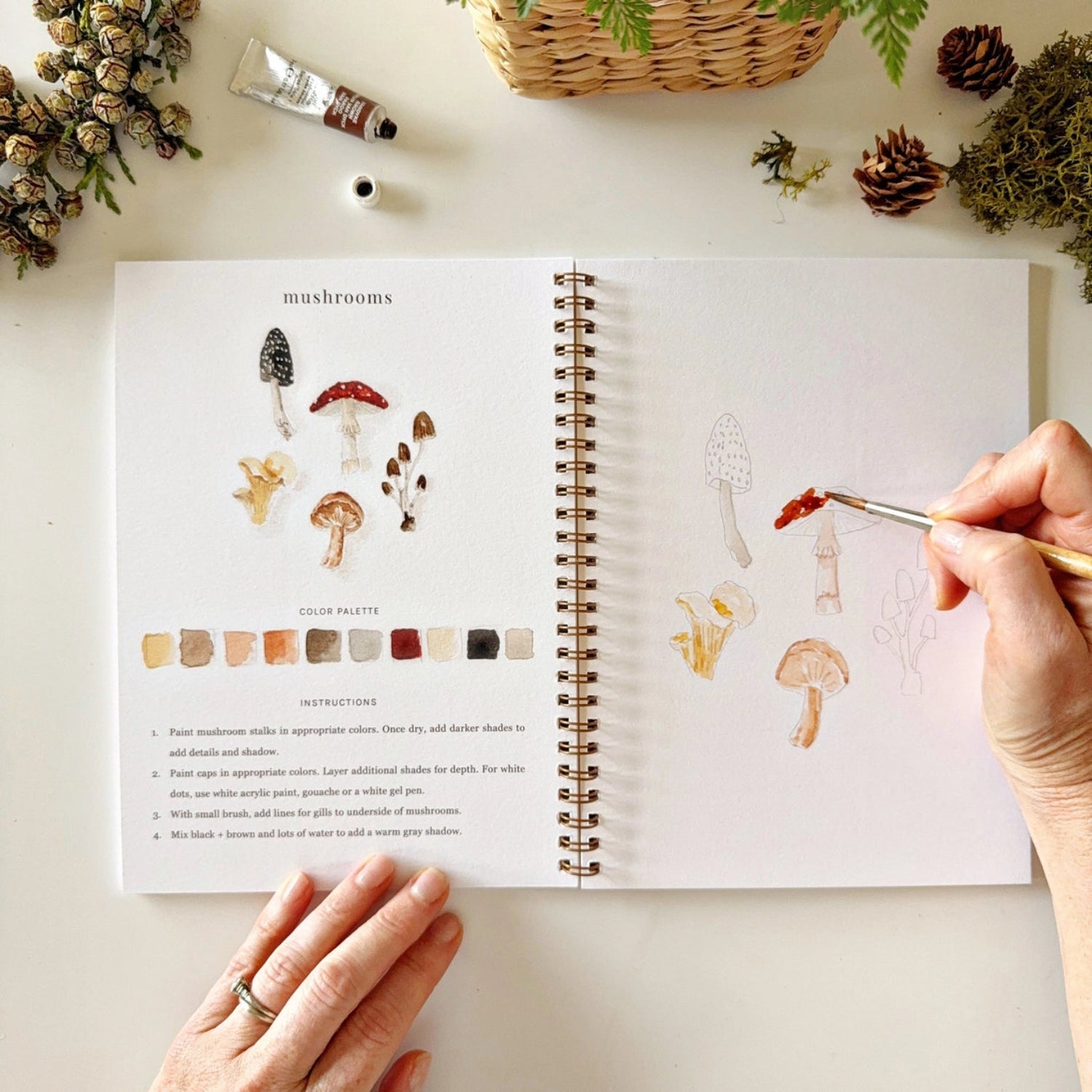 Open book with mushroom illustrations and a hand holding a paintbrush on a table with art supplies.