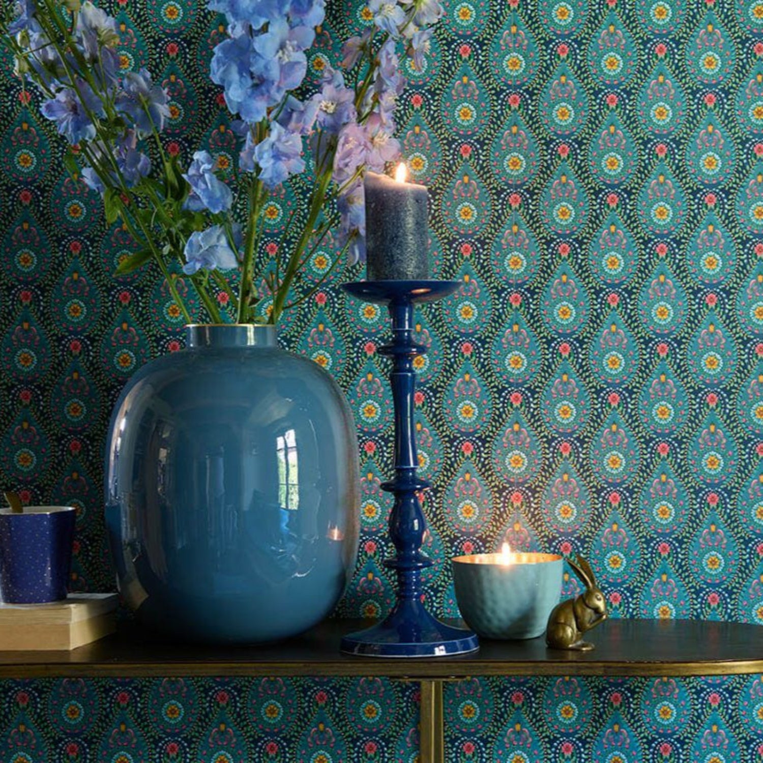 Decorative setting with a blue vase, candle, and flowers against a patterned wall.