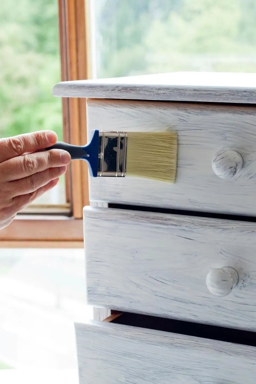 A person painting a dresser with white paint in whitewash style.