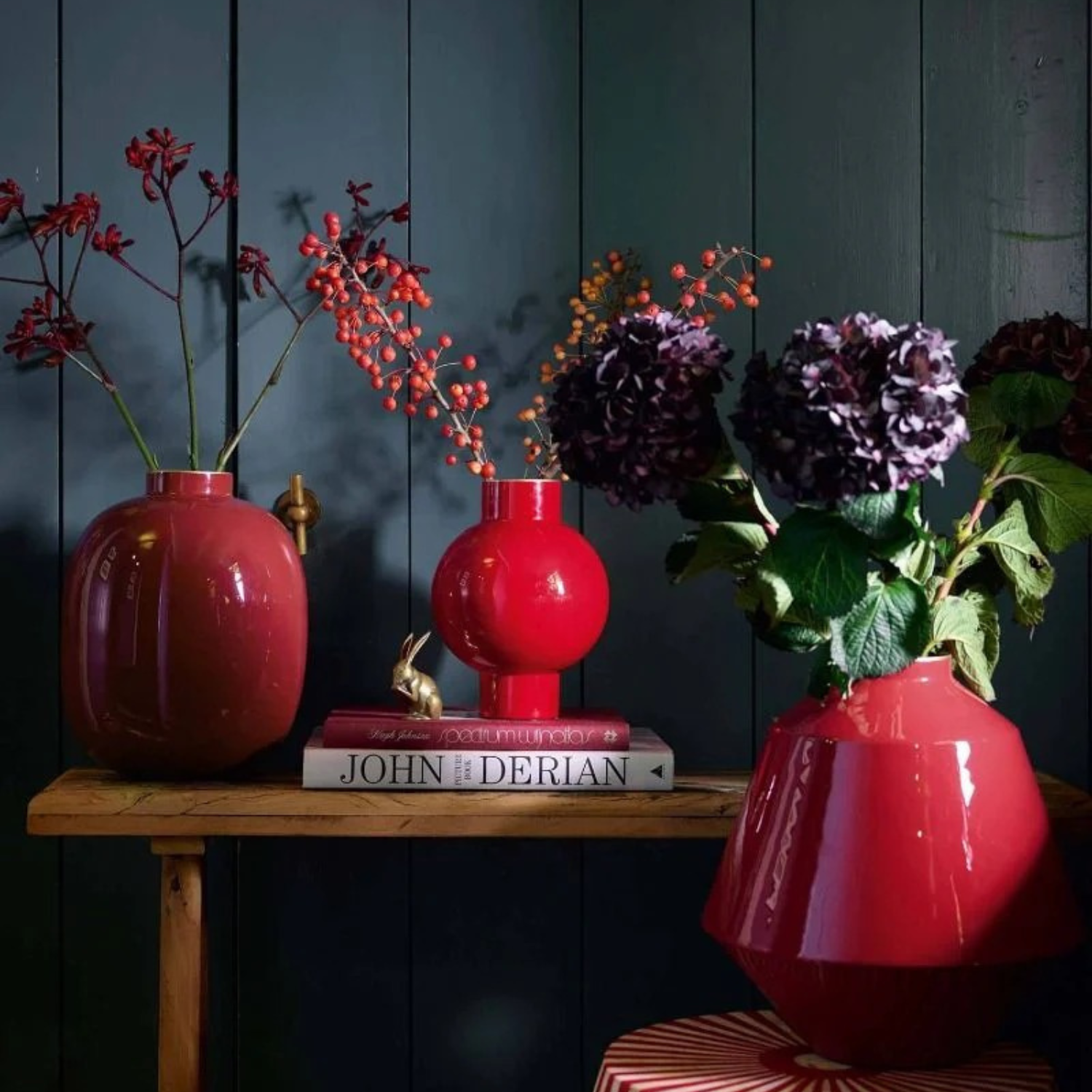 Three red vases with floral arrangements on a wooden table against a dark wooden panel wall.