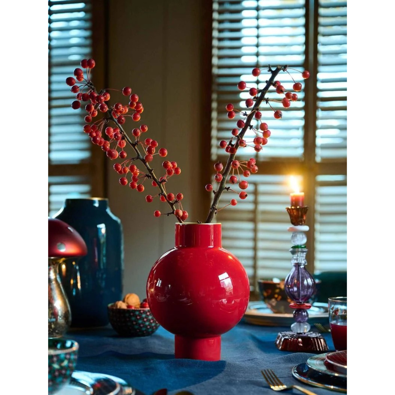 Red vase with branches on a table setting with candles and decor.