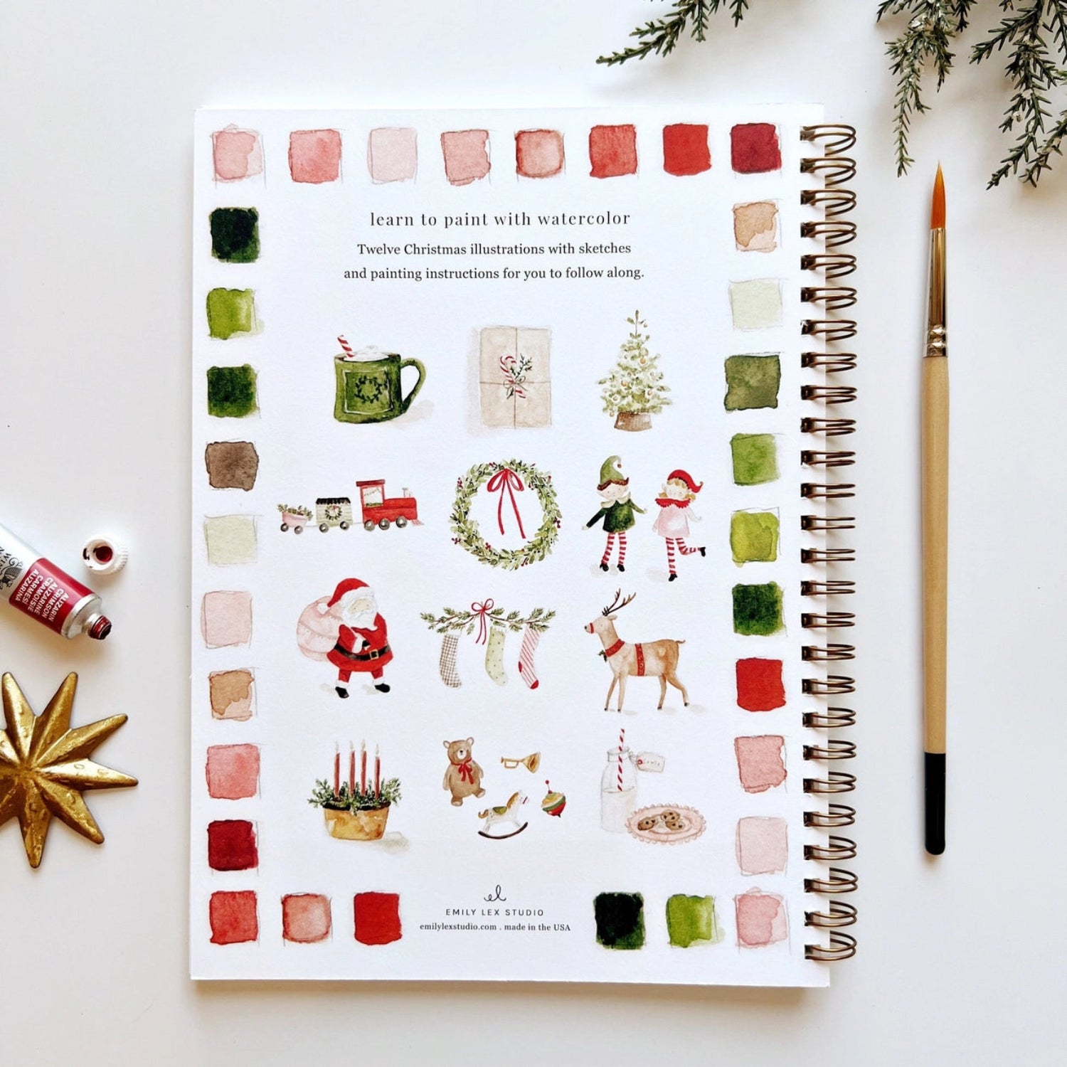 Spiral-bound art book with Christmas illustrations on a white surface with a paintbrush and decorative items.