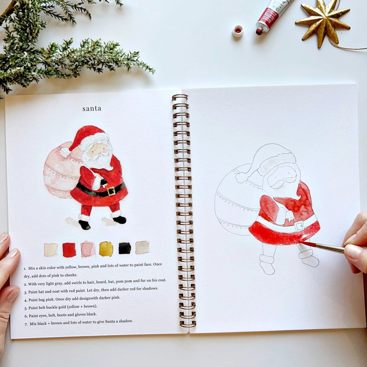 Open book with Santa Claus illustration and painting guide on a white surface with Christmas decorations.