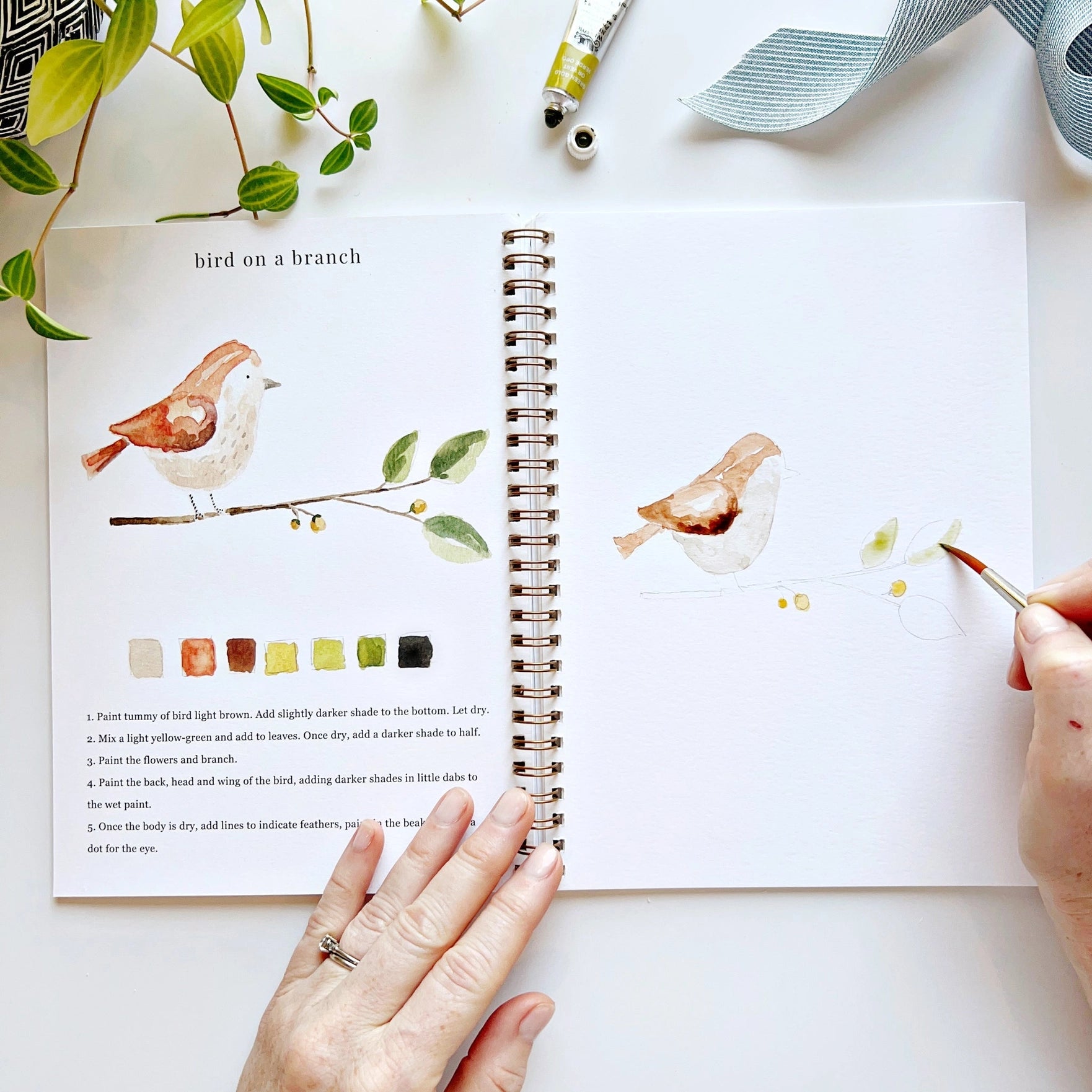 Person painting a bird on a branch with watercolors, using a book as a reference.