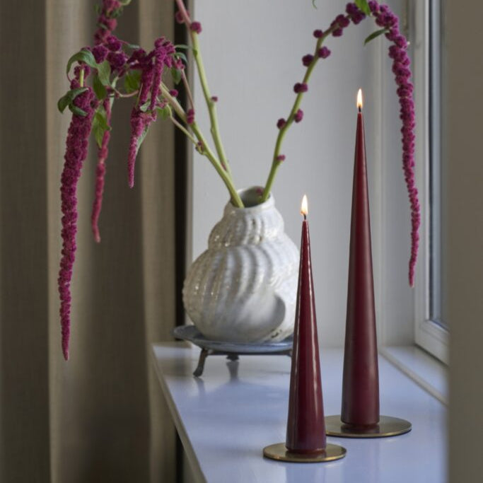 Two tall red candles on a windowsill with a white vase and pink flowers.