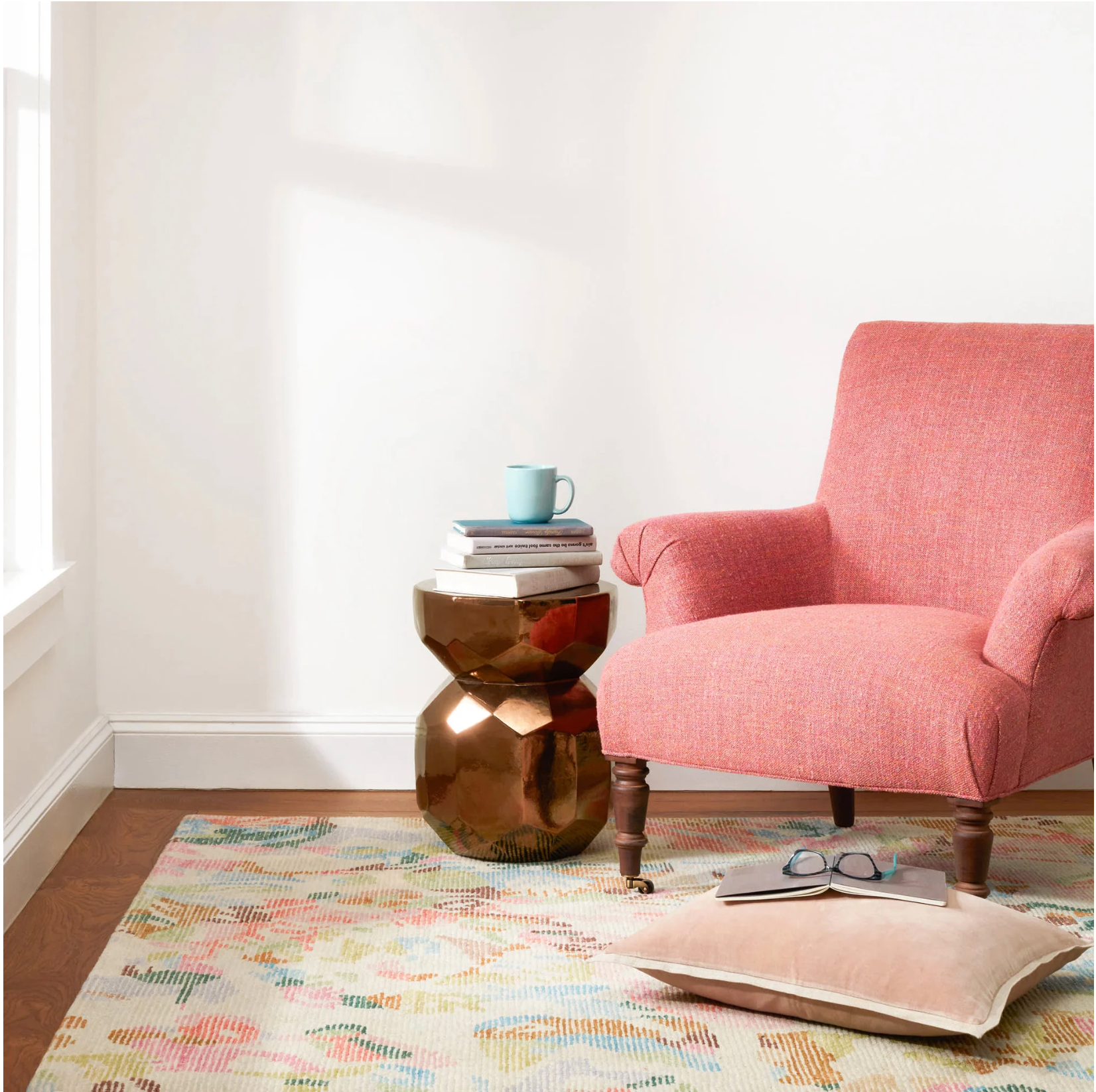 Pink armchair in a room with a gold side table and books on a rug.