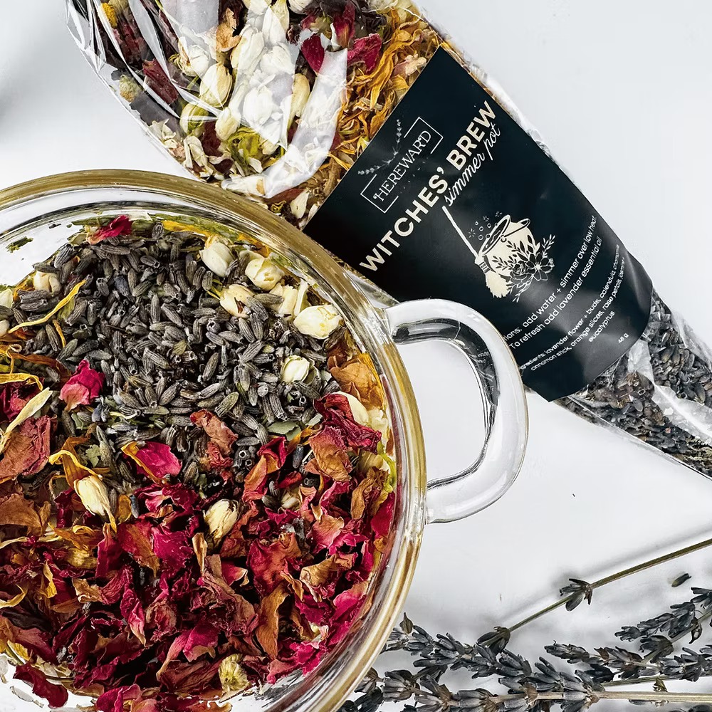 Glass teapot filled with dried herbs and flowers next to a package labeled 'Witches' Brew' on a white background.