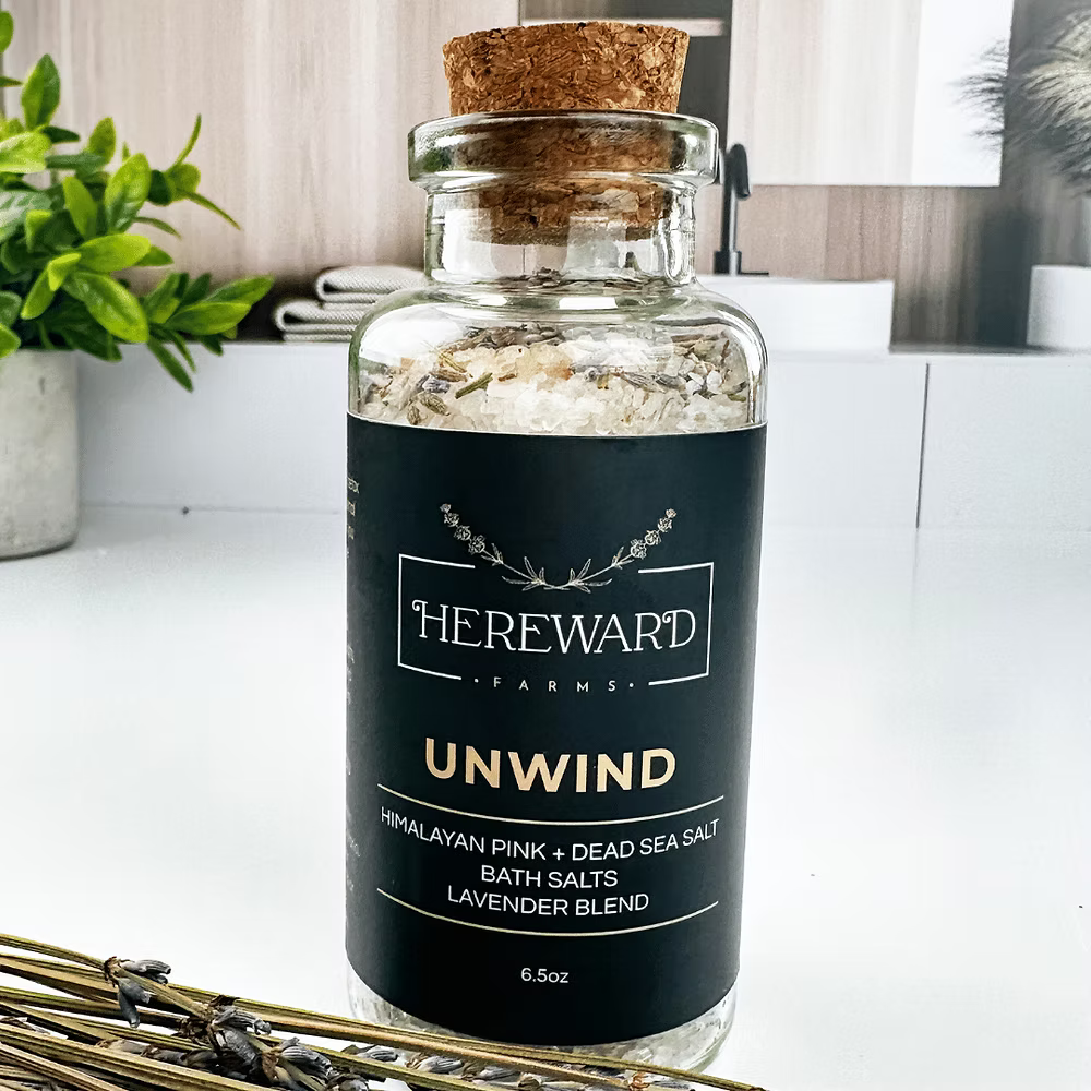 Glass jar of bath salts labeled 'Hereward Farms Unwind' on a white surface with plants.