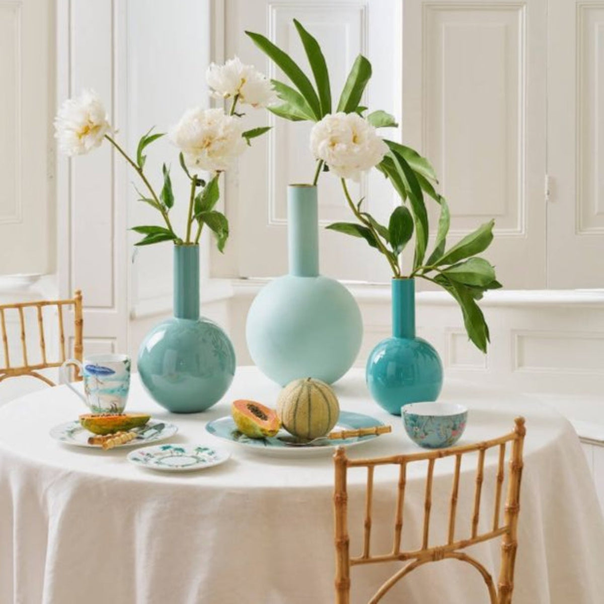 Dining table set with turquoise vases, white flowers, and fruit on a white tablecloth.