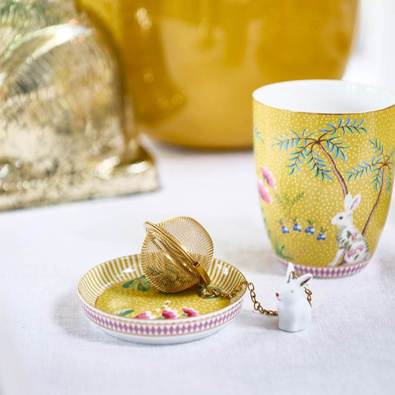 Yellow teacup with floral design on a white surface, accompanied by a small dish and tea ball.