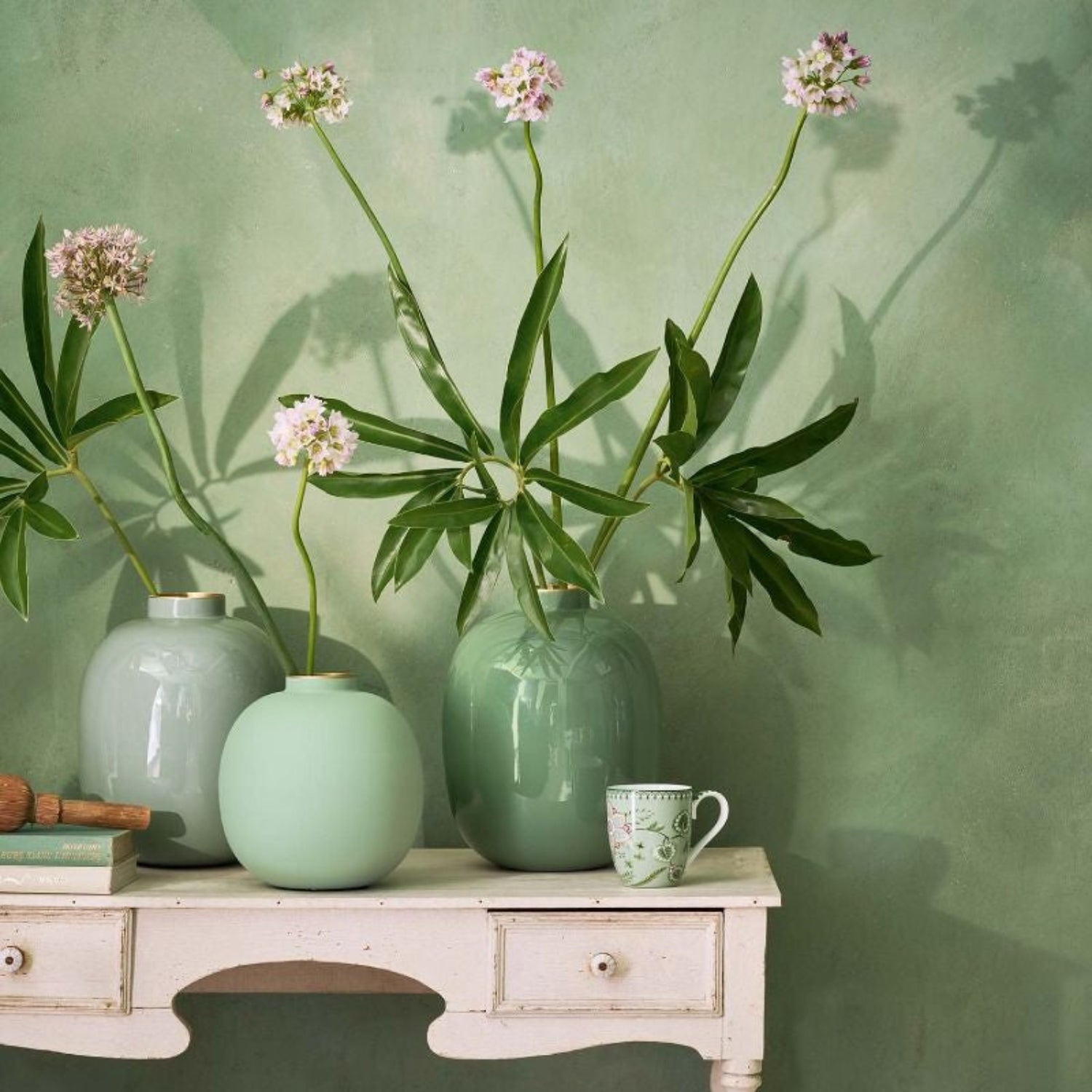 Green vases with flowers on a white table against a green wall